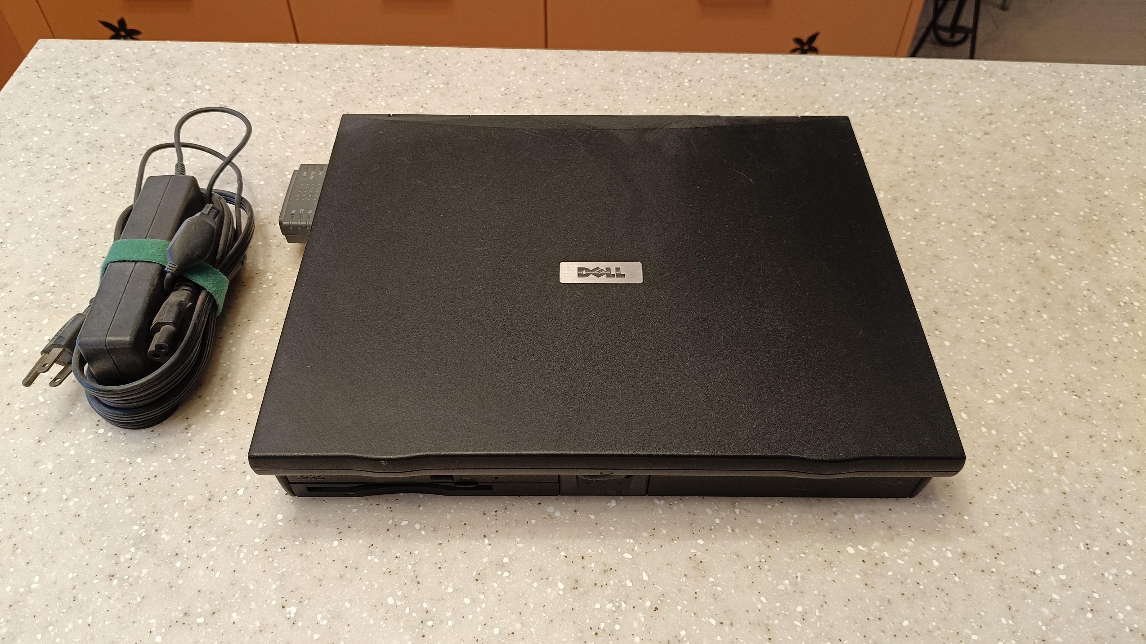 Old Dell laptop on kitchen counter booting up. Laptop is black and chunky 

Screen is closed. The charger is to the left of the image.