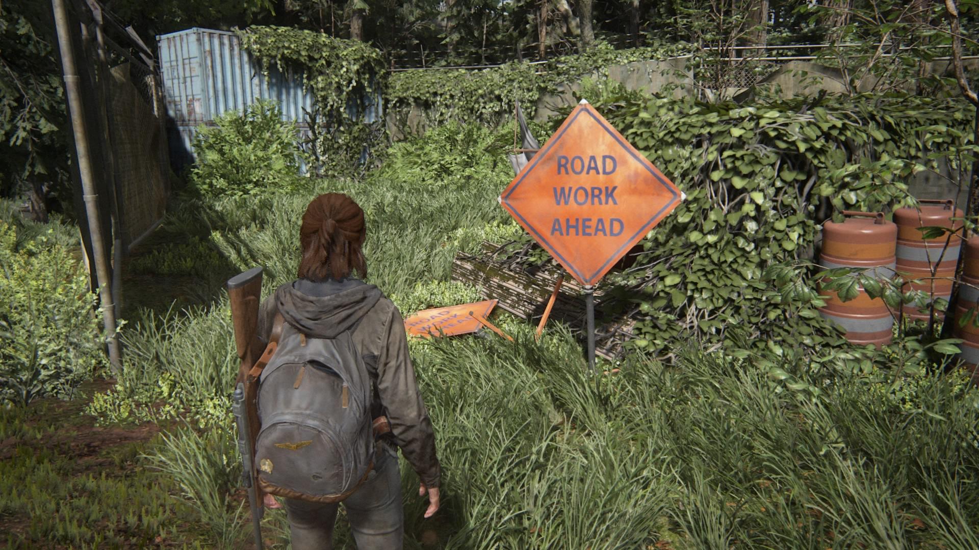 A screenshot from The Last of Us Part II. Ellie, a young woman in a shabby black hoodie and carrying an aged backpack and a rifle over her shoulder. She is standing on a long disused freeway, overgrown with vines. In front of her is an orange sign reading "Road work ahead"