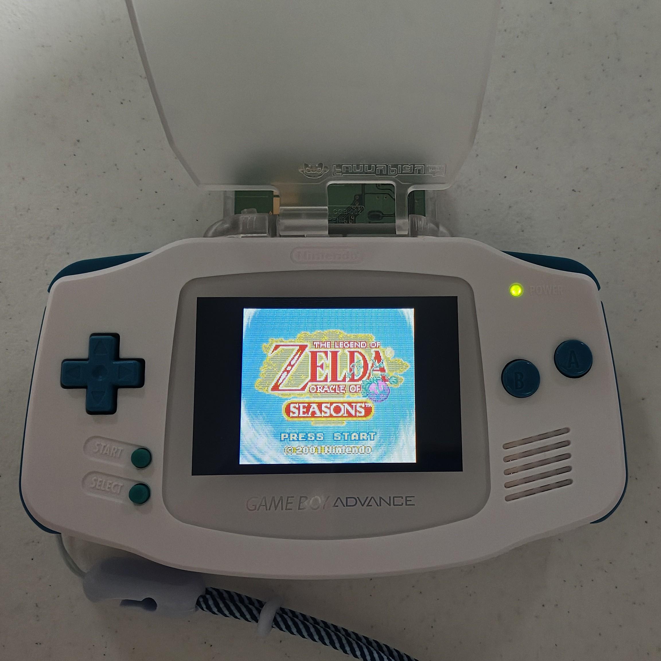 A white Game Boy Advance with a backlit screen showing the title screen for Legend of Zelda: Oracle of seasons.